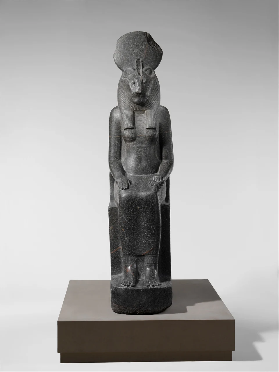 Sitzende dunkle Steinfigur mit Loewinnenkopf und Sonnenscheibe; die Haende ruhen auf den Knien.