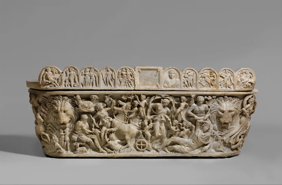 Breiter römischer Marmorsarkophag vor grauem Hintergrund mit dichtem Figurenrelief, einer gelagerten Person rechts, einem Wagen links und einer Inschrift auf dem Deckel.