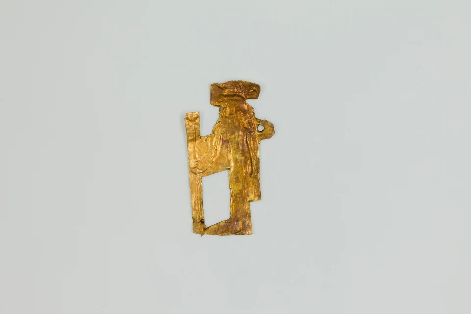 Kleines beschädigtes Goldblechamulett in menschlicher Silhouette auf hellgrauem Grund.