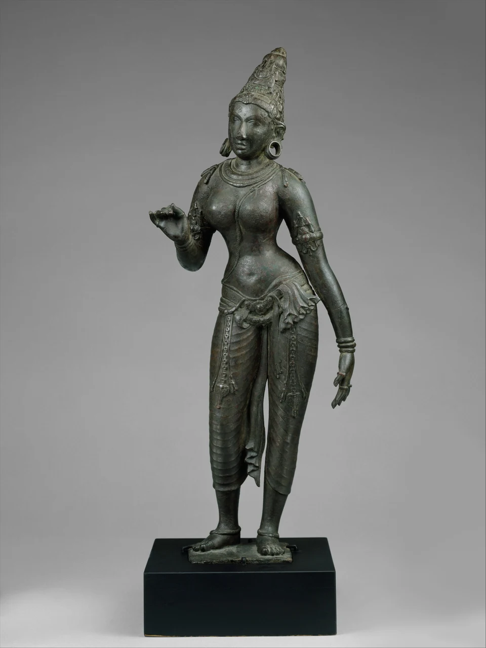 Grünlich patinierte stehende Metallfigur mit hoher Krone und Schmuck; ein Arm ist gebeugt, der andere hängt seitlich herab, das Gewand reicht bis zu den Unterschenkeln.