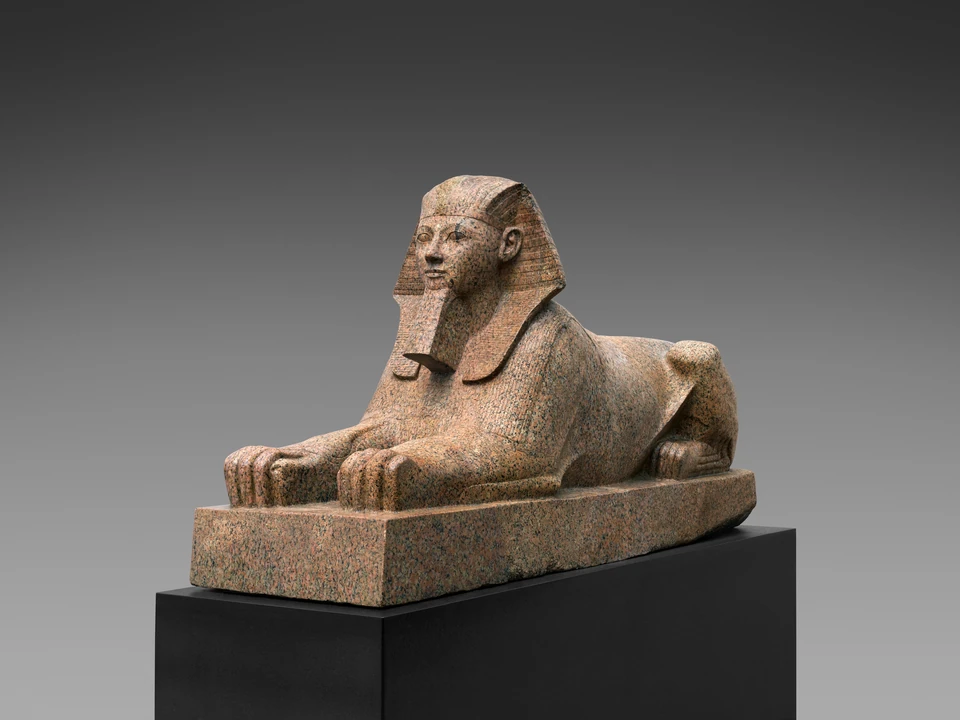 Rötliche Granitsphinx mit menschlichem Kopf, gestreiftem Kopftuch und Bart über einem ruhenden Löwenkörper. Die Vorderpfoten liegen nach vorn, die Hinterbeine sind eingezogen; Steinbasis vollständig sichtbar.