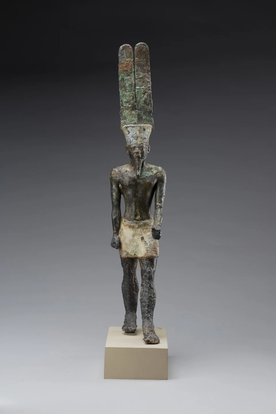Stehende dunkel patinierte Amun-Statuette mit zwei hohen Federn und hellem Schurz auf einem hellen Sockel.