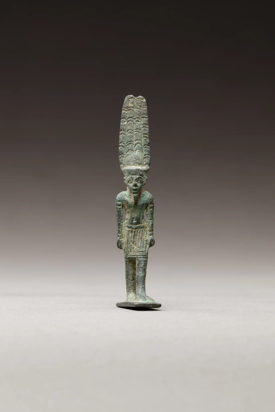 Kleine grünlich patinierte stehende Amun-Re-Figur mit hoher Federkrone vor dunklem Studiountergrund.