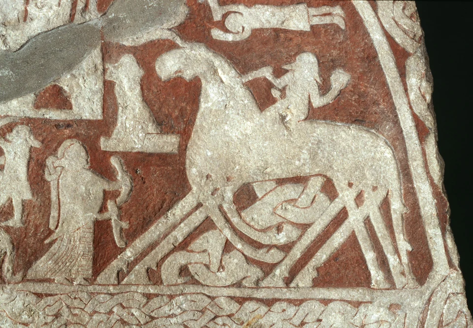 Fotografisches Detail eines Bildsteins: Reiter auf einem Pferd mit vielen parallelen Beinen, links eine Gestalt mit Horn. Rote Markierungen heben das Relief hervor; unten ein Flechtband.