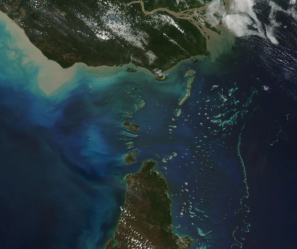 Satellitenaufnahme der Torres Strait: Zwischen zwei Küsten liegen Inseln und helle Riffe im blauen Meer; weiße Wolken ziehen über Land und Wasser.