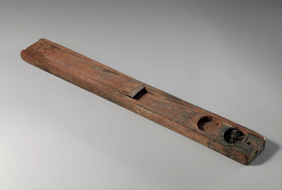 Lange hölzerne Schreiberpalette mit zwei runden Tintenmulden, eine davon dunkel gefüllt, vor grauem Grund.