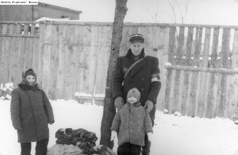 Zwei Kinder in Winterkleidung stehen neben Gepäck und einem Angehörigen der jüdischen Ghettopolizei im verschneiten Ghetto Łódź.