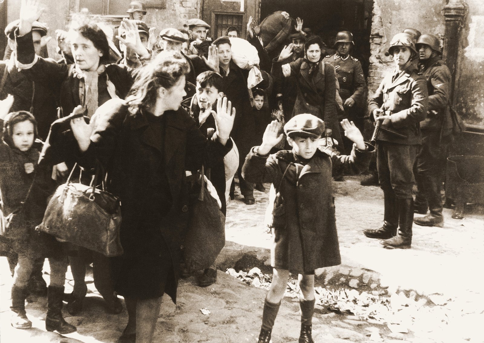 Jüdische Kinder und Erwachsene mit erhobenen Händen, bewacht von bewaffneten Männern im Warschauer Ghetto, 1943.