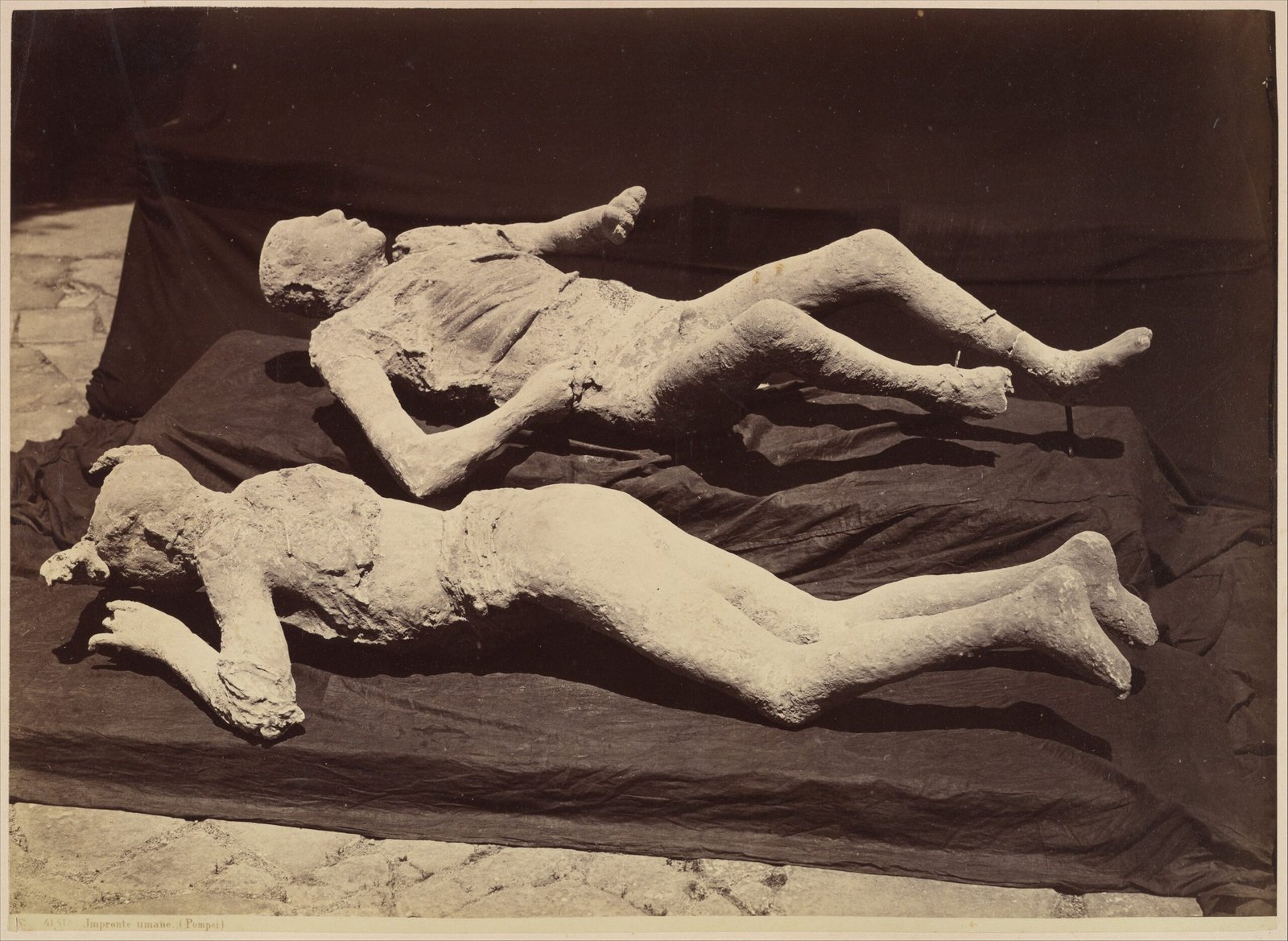 Historische Fotografie zweier Gipsabgüsse von Opfern aus Pompeji auf einem dunklen Stoffuntergrund; beide liegend, einer mit angewinkelten Knien.